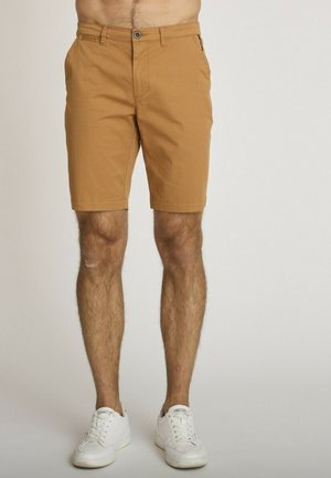 Homme portant un short beige au genou et des baskets blanches à lacets, debout devant un fond uni.
