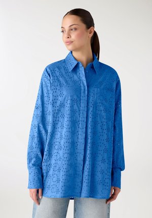 Donna che indossa una camicia azzurra larga con occhielli, maniche lunghe e colletto, abbinata a jeans chiari, in piedi davanti a uno sfondo semplice.