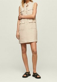 Donna che indossa un vestito senza maniche in lino beige con tasche frontali e sandali neri aperti, in piedi contro uno sfondo chiaro e semplice.
