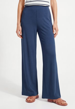 Kobieta ubrana w luźne granatowe spodnie, w połączeniu z beżowymi sandałami na odkryte palce i pasiastą bluzką, stojąca na białym tle.