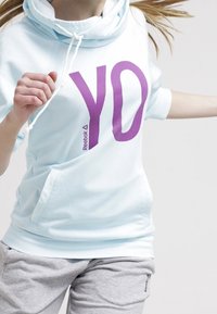 Sudadera azul claro con una gran impresión morada de "YO" y capucha con cordón. Combinada con pantalones de chándal grises que presentan un logo.