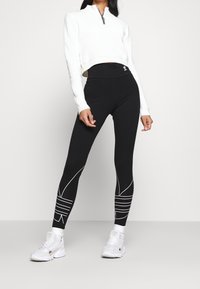 Svarta leggings med vita linjemönster och hög midja, kombinerat med en kortärmad vit tröja med dragkedja halvvägs. Vita sneakers.
