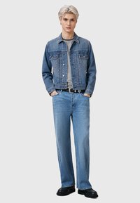 Jeans jakna u srednje plavoj boji s dva džepa na prsima, uparena s izblijedjelim plavim hlačama širokih nogavica i crnim robustnim cipelama. Ima prugastu majicu ispod.