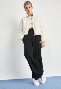 Puffig krämfärgad bomberjacka med dragkedja, svarta vida byxor med vitt Adidas-logotyp, och vita sneakers med accentstrimmor.