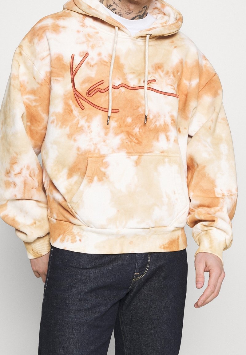 En tie-dye hoodie i nyanser av orange och grädde, med en frontficka, dragsko och broderad logga i rött, tillverkad av mjukt tyg.