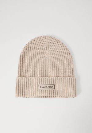 Beige neulepipo, jossa on ribbikuviointi ja taitettu reunus. "Calvin Klein" -merkkilappu on ommeltuna suorakaiteen muotoiseen palaseen.