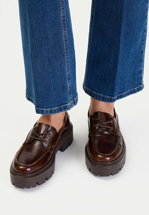 Mocasines de plataforma de cuero marrón usados con vaqueros azules de pernera recta sobre un fondo liso y claro.