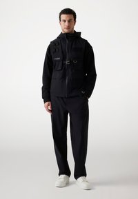 Chaqueta negra con múltiples bolsillos frontales, cierre de cremallera y correas ajustables. Combinada con pantalones negros y zapatillas blancas.