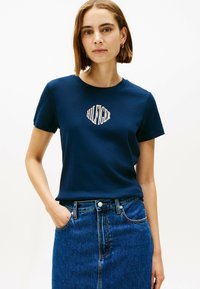 T-shirt in cotone blu navy con un logo grafico centrale in bianco. Maniche corte e scollatura a girocollo. Abbinato a una gonna di jeans.