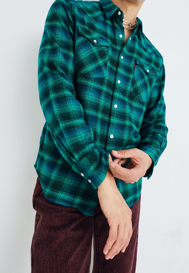 Camisa de franela a cuadros verdes y azules con cuello abotonado, bolsillos en el pecho y botones plateados; combinada con pantalones burdeos.