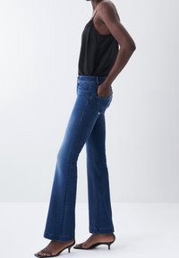 Jean bleu coupe slim avec légère ouverture au bas, porté par une personne en haut noir sans manches et sandales à talons ouvertes noires, debout avec une main dans la poche.