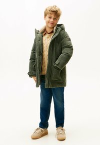 Grøn velourjakke med foret hætte, to forlommer og lynlås_lukning. Bæres over en beige strikket sweater og blå jeans.