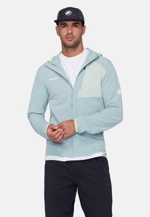 Uomo che indossa una giacca Mammut azzurro chiaro con cerniera, t-shirt bianca, cappello nero con logo e pantaloni neri, in piedi davanti a uno sfondo semplice.