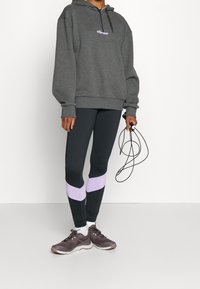 Harmaa huppari, jossa logo, yhdistetty mustiin leggingseihin, joissa on violetit raidat; malli pitelee kuntohyppynarua; urheilukengät.