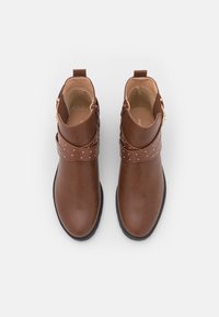 Hnědé kožené kotníkové boty s kulatou špičkou, ozdobným páskem se zlatými cvoky a bočním zipem pro snadné obouvání.