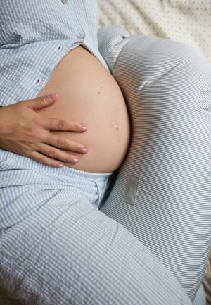 Grūtnieču spilvens ar svītrainu audumu, izliekta forma atbalstam; roka atpūšas uz noapaļotas grūtnieces vēderiņa, mīksta tekstūra, gaišas krāsas.