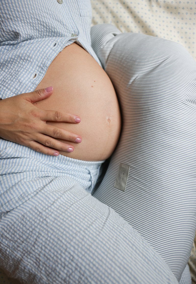 Travesseiro de maternidade com tecido riscado, forma curva para suporte; mão repousada sobre uma barriga de grávida arredondada, textura suave, cores claras.