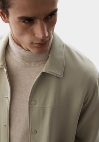 Chaqueta de botones verde claro con cuello, llevada sobre un suéter de cuello alto beige. Tejido suave, diseño sencillo, sin patrones visibles.