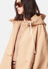 Beige Kapuzenjacke mit glatter Textur, verfügt über einen Front-Reißverschluss, Kordelzüge und aufgesetzte Taschen. Über einem strukturierten hellrosa Oberteil getragen.