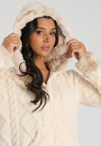 Cremefarvet hættetrøje i fleece med et tekstureret kabelstrik mønster. Har blød faux fur-foring i hætten og ved håndleddene. Lynlåslukning.