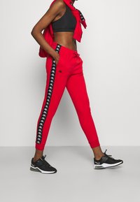 Rode joggingsbroek met elastische boorden en een trekkoord in de taille, met zwarte zijstrepen met een herhaald logo-patroon. Gecombineerd met zwarte sneakers.