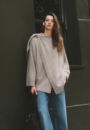 Kvinde med langt hår iført en løs beige sweater og blå jeans står op ad en mørk væg og holder en sort taske i den ene hånd.