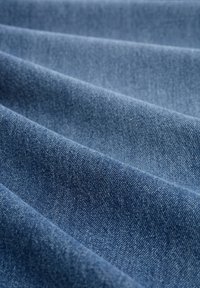 Tejido de denim en un color azul medio con una textura suave y un patrón de tejido visible, mostrado en pliegues para resaltar su grosor y flexibilidad.