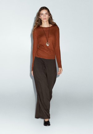 Brown long-sleeve top with a round neckline paired with loose, dark brown pants. Pendant necklace adds a metallic accent.