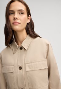 Giacca camicia beige con colletto, due tasche sul petto e chiusure a bottone marrone. Texture liscia con dettagli di cucitura visibili.