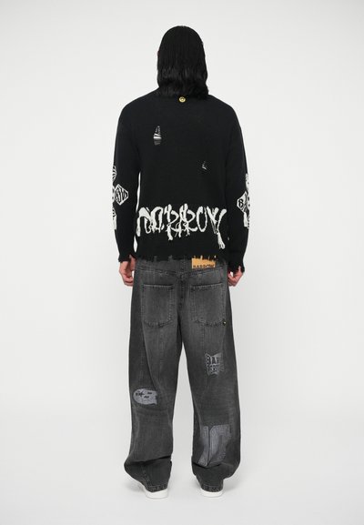 Maglione nero a maglia con grafiche bianche sulle maniche e sull'orlo, indossato con jeans grigi larghi caratterizzati da toppe e cuciture in vista.