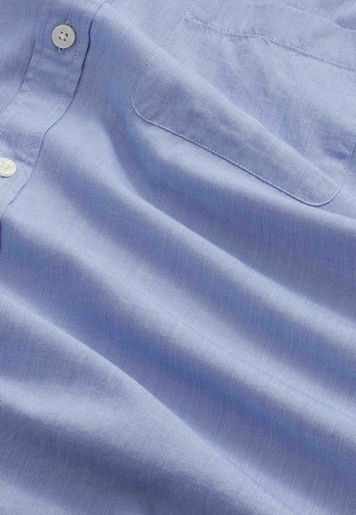 Tessuto di camicia di cotone azzurro chiaro con bottoni bianchi e una tasca sul petto cucita, che mostra la texture e leggere pieghe.