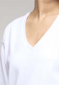 Primer plano del cuello y la parte superior del pecho de una persona que lleva una camiseta blanca lisa con cuello en V contra un fondo neutro.