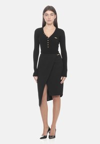 Schwarzes, geripptes Oberteil mit goldenen Knöpfen, kombiniert mit einem schwarzen Wickelrock mit hohem Schlitz, akzentuiert durch ein Detail mit goldener Schnalle.