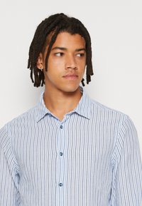 Camisa de botones a rayas en azul claro y blanco, con cuello, mangas largas y botones azules. Material de algodón con una textura suave.