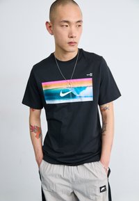 Svart Nike T-skjorte med et fargerikt grafisk motiv av bølger. Korte ermer, rund hals, mykt bomullsmateriale. Modellen har på seg lyse bukser.