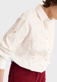 Camicia bianca in cotone con fiori ricamati dettagliati sulle maniche e chiusura con bottoni. Indossata con pantaloni di velluto a coste bordeaux.