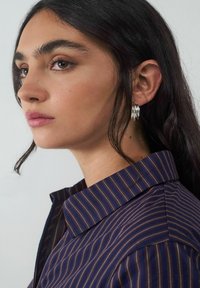 Marineblaue Bluse mit hellbraunen vertikalen Streifen, mit Kragen, kombiniert mit silbernen Creolen. Glatte Stoffstruktur.