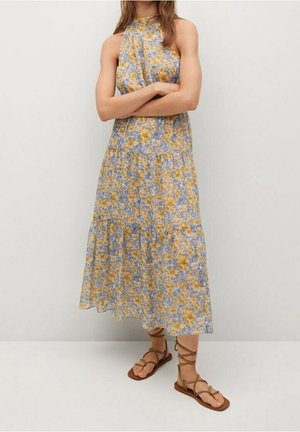 Kobieta w sukience bez rękawów, z wysokim kołnierzem, sięgającej kostek, w kwiatowy wzór w odcieniach żółci, niebieskiego i beżu, nosząca brązowe sandały na płaskiej podeszwie z wiązaniami, stojąca z założonymi ramionami.