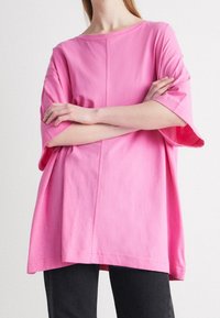 Camiseta de algodón rosa de gran tamaño con mangas anchas, cuello redondo y un detalle de costura central, combinada con pantalones negros.