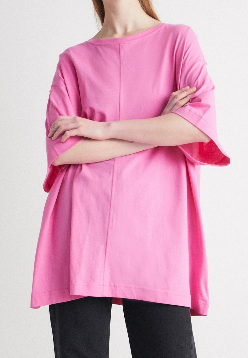Camiseta de algodón rosa de gran tamaño con mangas anchas, cuello redondo y un detalle de costura central, combinada con pantalones negros.