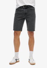 Shorts en denim noir avec une coupe décontractée, dotés de passants de ceinture et de coutures visibles. Portés avec des baskets blanches et des chaussettes blanches mi-mollet.