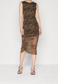 Vestido marrón hasta la rodilla con un patrón floral, diseño sin mangas y detalle fruncido en el costado. Combinado con sandalias negras de tiras.