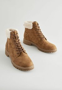 Botas de tobillo de ante marrón con un suave puño de piel blanca, diseño con cordones y suela de goma texturizada con tacos pronunciados para mayor tracción.