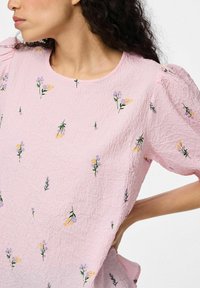 Top rosa, testurizzato, con maniche a sbuffo, che presenta motivi floreali ricamati in giallo e viola su uno sfondo stropicciato.