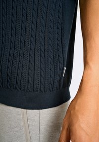 Granatowy sweter dzianinowy z teksturowanym wzorem w warkocze. Posiada ściągacz na dole i małą metkę z logo. Szare spodnie widoczne na dole.