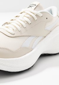 Beige sportskor med en texturerad mesh-övre, släta läderdetaljer, vit randdetalj och en vadderad vit sula med svart yttersula.