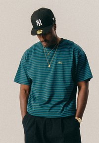 Férfi fekete New York Yankees sapkát visel, kék és fekete csíkos pólóban, arany nyaklánccal és órával, keze a nadrágzsebében van és lefelé néz.