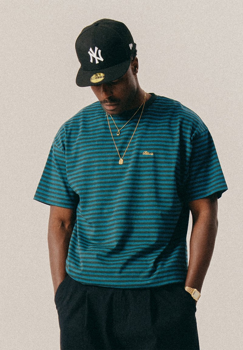 Férfi fekete New York Yankees sapkát visel, kék és fekete csíkos pólóban, arany nyaklánccal és órával, keze a nadrágzsebében van és lefelé néz.