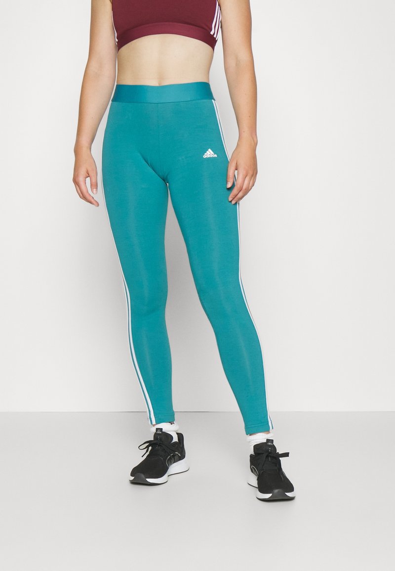 Kvinna iförd teal Adidas leggings med vita ränder, svarta sneakers och en vinröd sport-bh, stående mot en enkel ljus bakgrund.