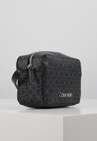 Czarna prostokątna torebka wykonana z fakturowanego materiału z powtarzającym się wzorem logo. Srebrny zamek i akcent z nazwą marki wytłoczoną z przodu.